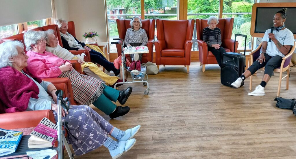 A group of elderly women sit in a bright lounge, enjoying fun activities such as balloon tennis while a man with a microphone speaks. Some use walking frames, and books and knitting items can be seen.