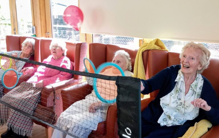 Four elderly women seated in chairs enjoy a lively game of balloon tennis with racquets over a small net, improving their flexibility in a bright, sunlit room.
