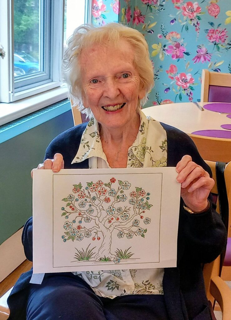 An elderly woman sits indoors, smiling and holding up a colourful drawing of a tree with flowers on its branches, possibly a delightful present from young visitors.