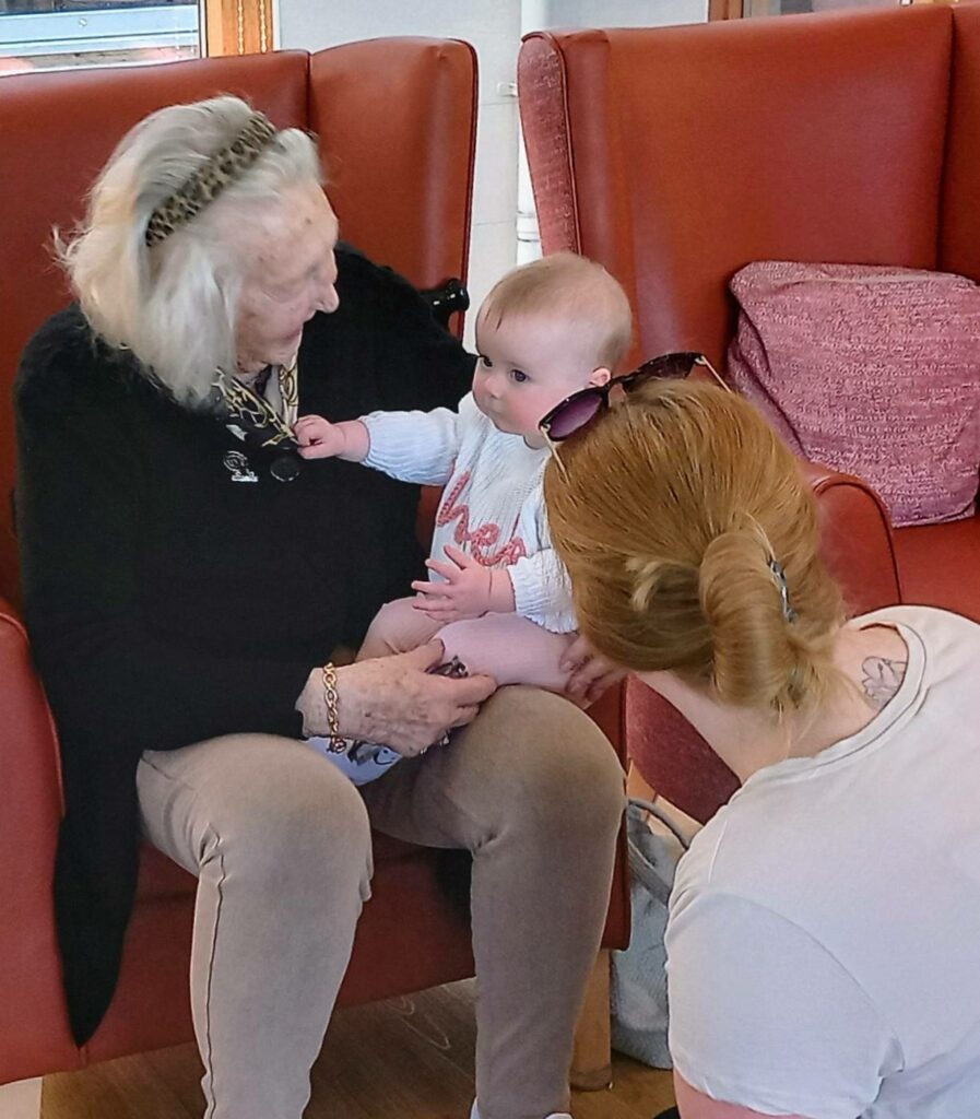 At Lear House, generations connect as an elderly woman cradles a baby on her lap, whilst a red-haired woman crouches nearby, sharing in the warmth of the moment.