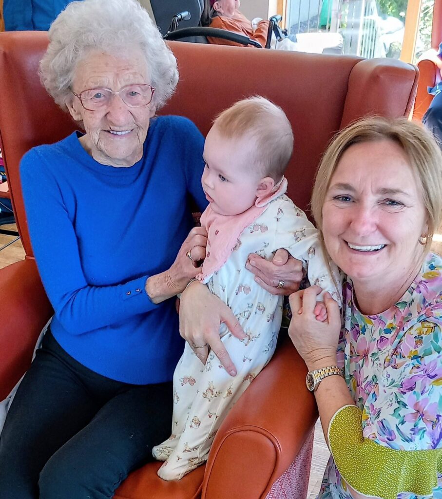 An elderly woman sits in a red armchair at Lear House, holding a baby, while another woman kneels beside them, sharing intergenerational smiles for the camera.
