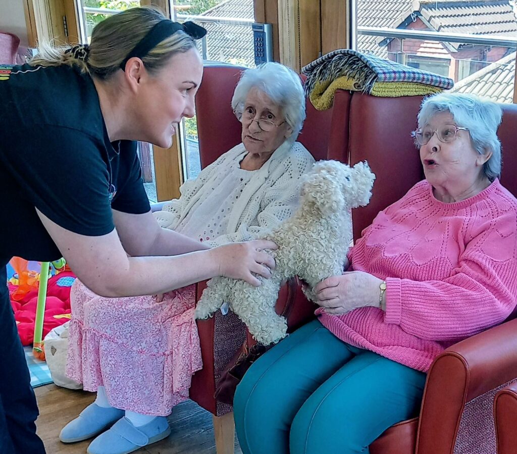 At Lear House, cheerful faces from different generations are seen as a woman holds a small, fluffy dog beside two elderly women in armchairs; one softly strokes the dog while the other observes.