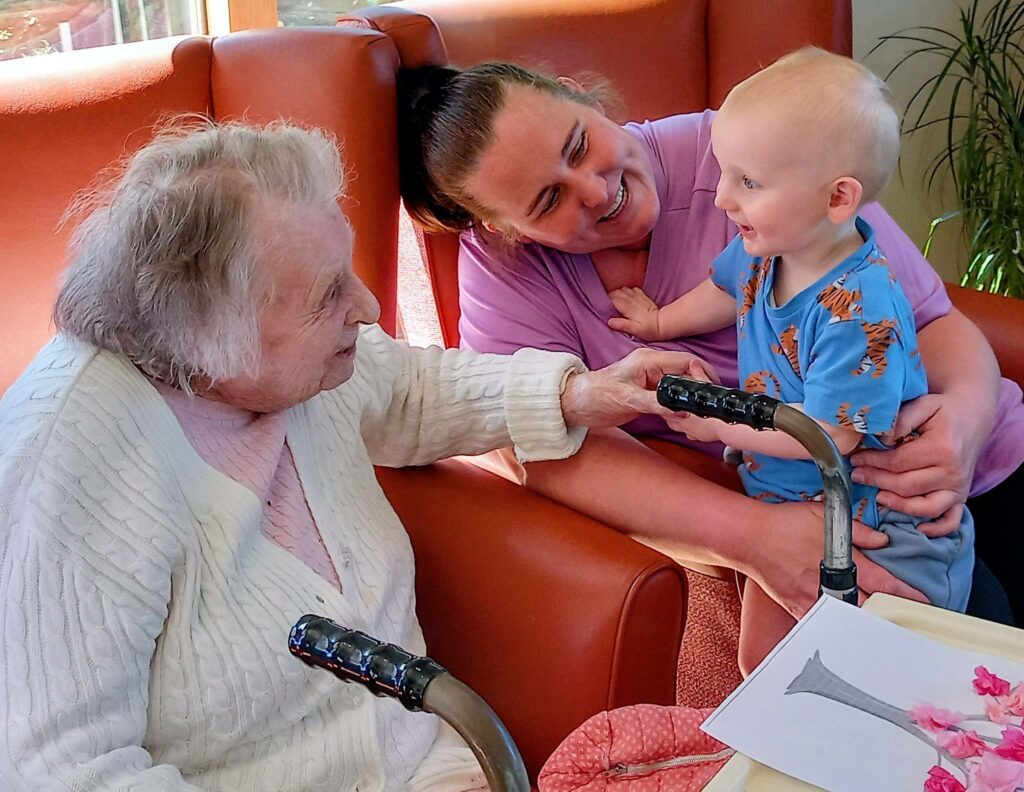An elderly woman in a white jumper shares intergenerational smiles as she holds hands with a baby on a woman's lap, all seated in the brightly lit Lear House.