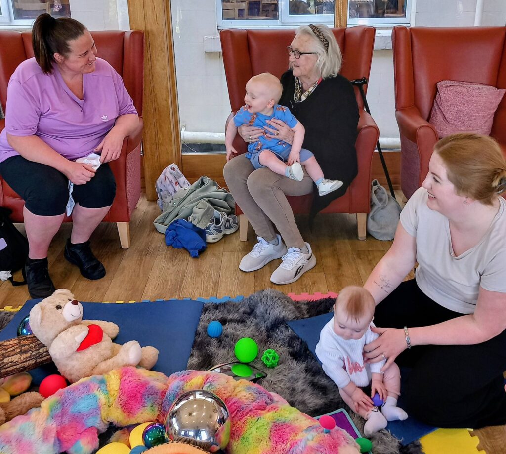 At Lear House, intergenerational smiles fill the room as two women sit in armchairs holding babies, while another woman sits on the floor with a baby among toys and colourful blankets.