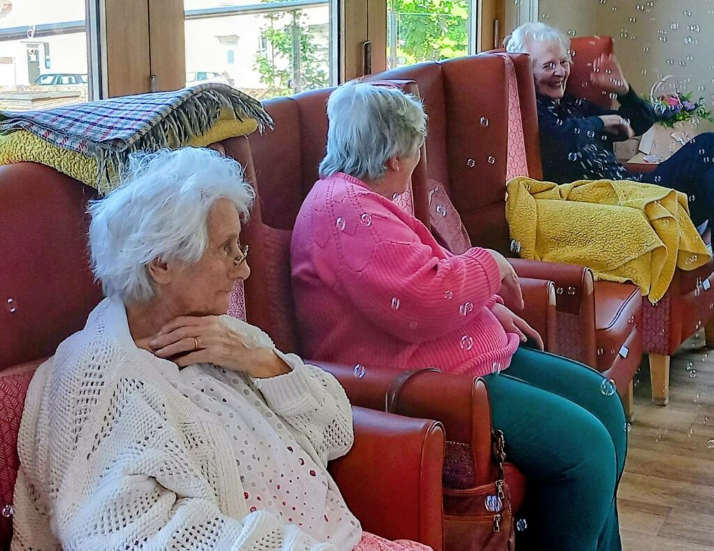 At Lear House, three elderly women sit on red armchairs in a bright room, surrounded by floating bubbles and intergenerational smiles, with cosy blankets draped over some of the chairs.