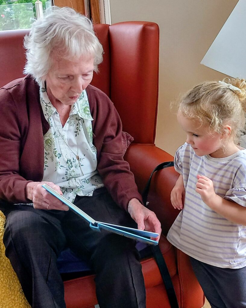 An elderly woman at Lear House sits on a red chair, reading to a young girl next to her—an intimate moment filled with intergenerational smiles as they concentrate on the book together.