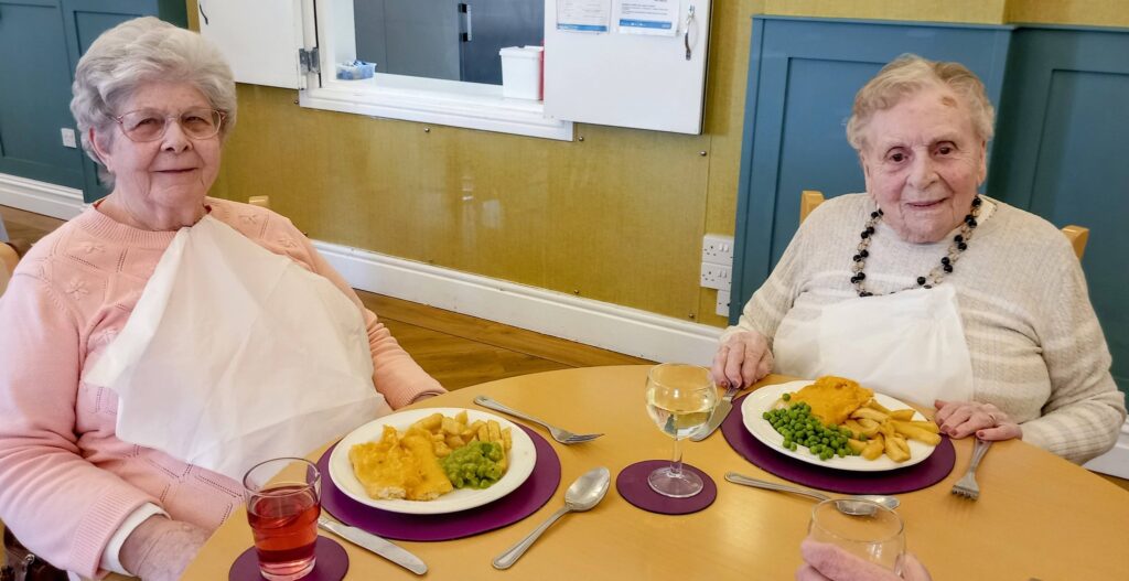 Two elderly women sit at a table with plates of fish, chips and peas, along with drinks in glasses. Both are wearing pale jumpers and serviettes, seated in a dining area.
