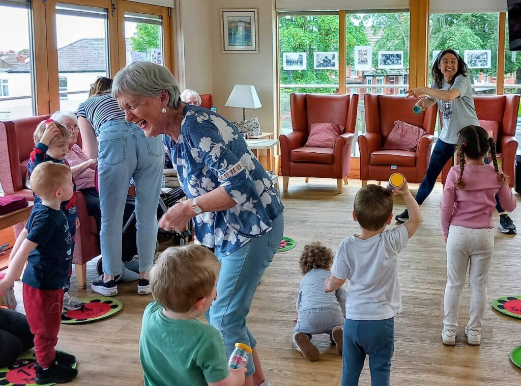 Older adults and young children share smiles as they enjoy intergenerational play together in a bright Lear House room filled with large windows, chairs, and playful activities.