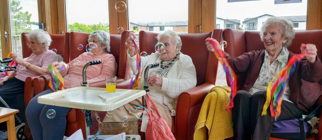 Four elderly women sit in armchairs at Lear House, holding colourful ribbons and sharing smiles, as bubbles drift around them in a bright communal lounge.