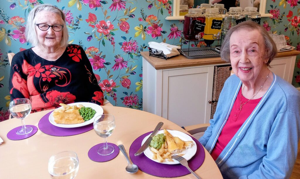 Two elderly women sit at a table with fish, chips, peas, and glasses of water in front of them. The background features floral wallpaper and dispensers with drinks.