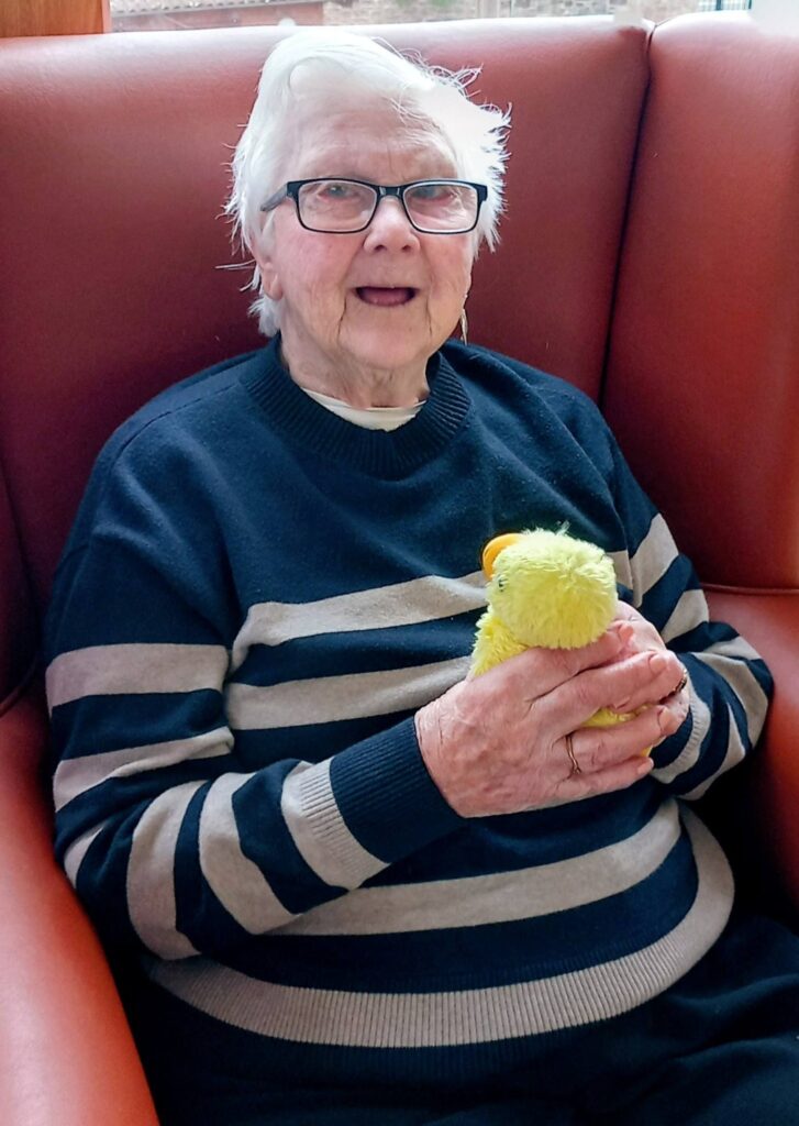 Elderly woman wearing glasses and a striped jumper sits on a red armchair at Lear House, smiling warmly and holding a small yellow cuddly duck.