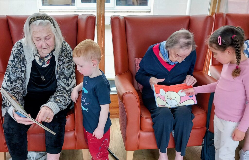 At Lear House, two elderly women and two young children share smiles as each pair enjoys a picture book. The children stand beside the women seated in armchairs—an intergenerational moment.