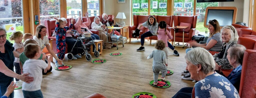 Children and elderly people share intergenerational smiles as they interact and play together in a brightly lit Lear House common room with large windows, red chairs, and colourful mats.