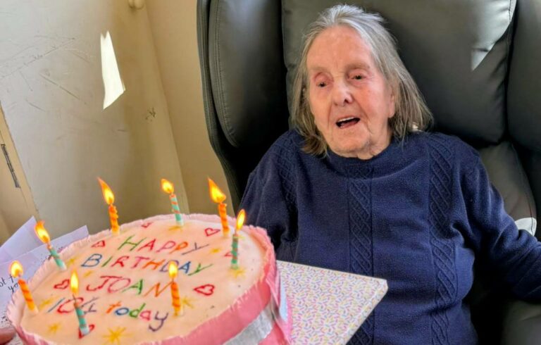 An elderly woman sits in a chair, looking at a pink birthday cake with lit candles and 'Happy Birthday Joan' written on it, held by another person.