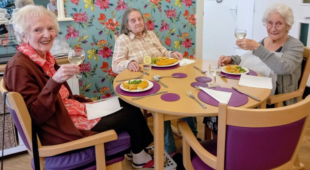 Three elderly women sit around a circular table with plated meals and glasses of white wine, lifting their glasses in a joyful toast. Floral wallpaper adorns the background.