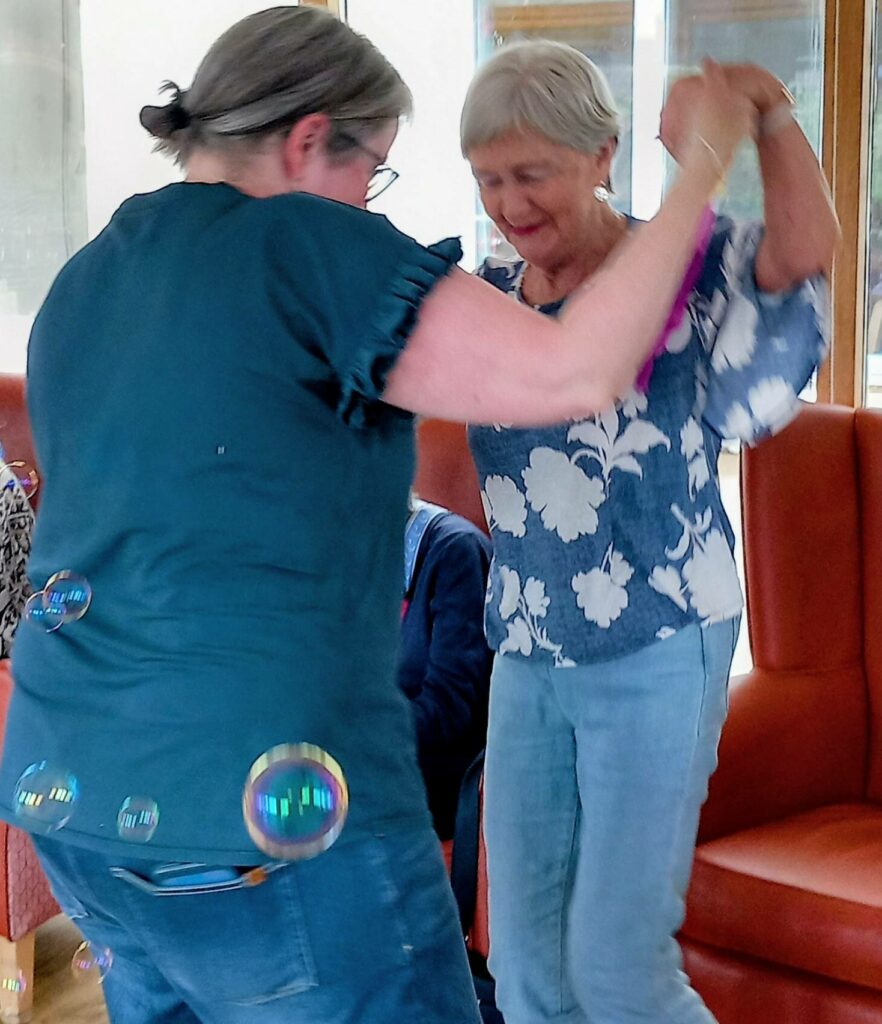 Two older women are holding hands and dancing indoors at Lear House, smiling as bubbles drift around them.