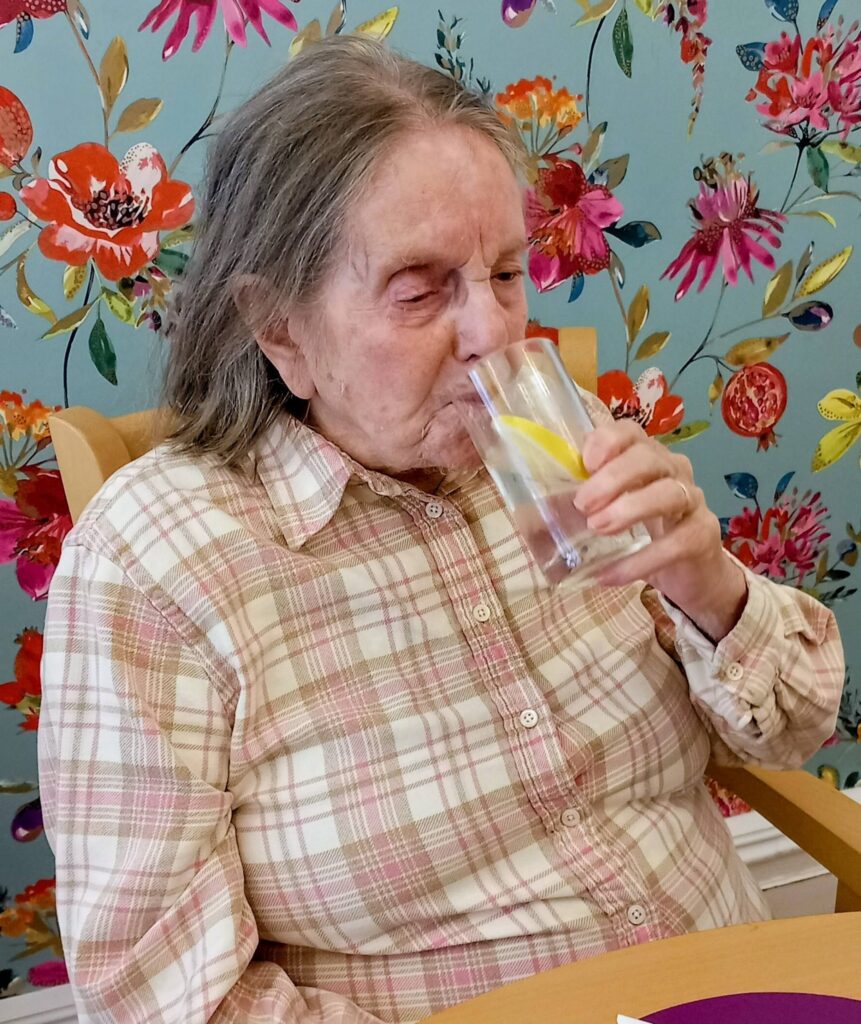 An older woman in a checked shirt sits at a table and sips a glass of water with a slice of lemon, set against a backdrop of colourful floral wallpaper.