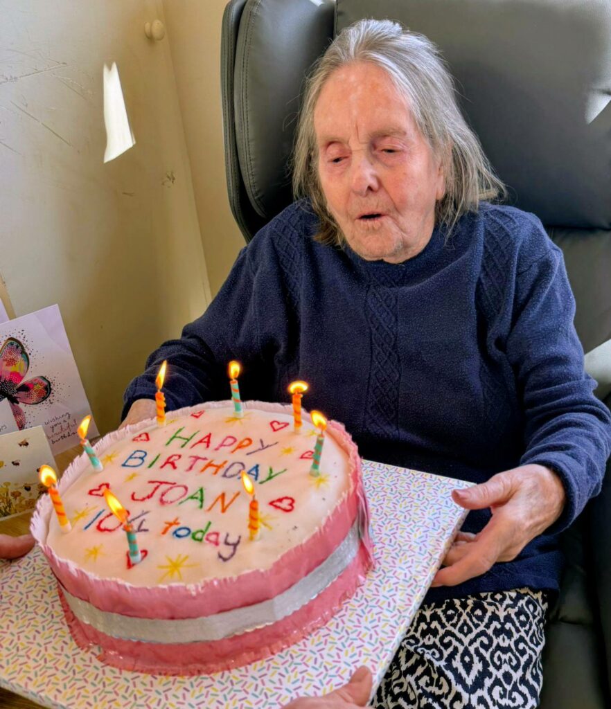 Elderly woman seated in a chair holds a pink-iced birthday cake with lit candles and "Happy Birthday Joan 101 today" written on it.