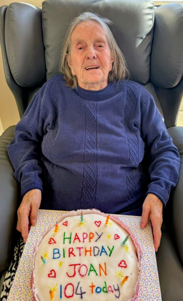 An elderly woman sits in an armchair holding a decorated birthday cake that reads Happy Birthday Joan 104 today.