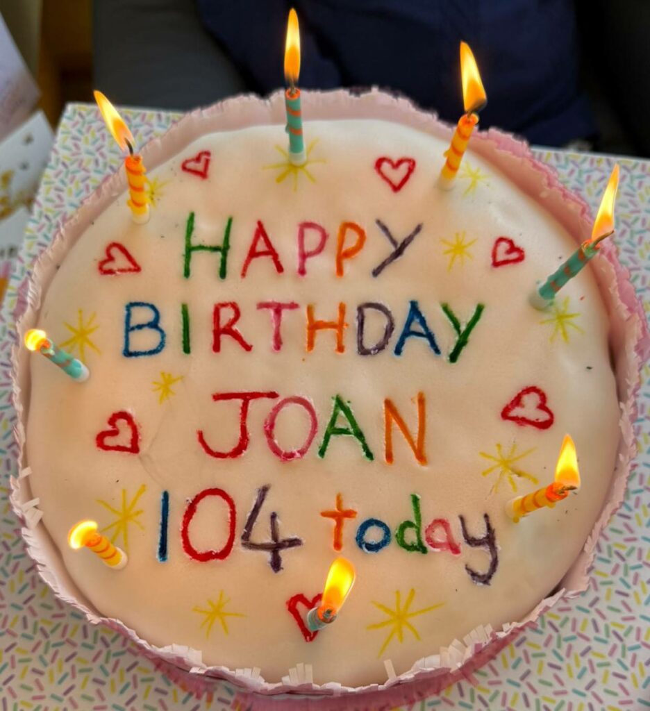 A birthday cake with lit candles and the message ‘Happy Birthday Joan 104 today’ written in colourful icing on top.