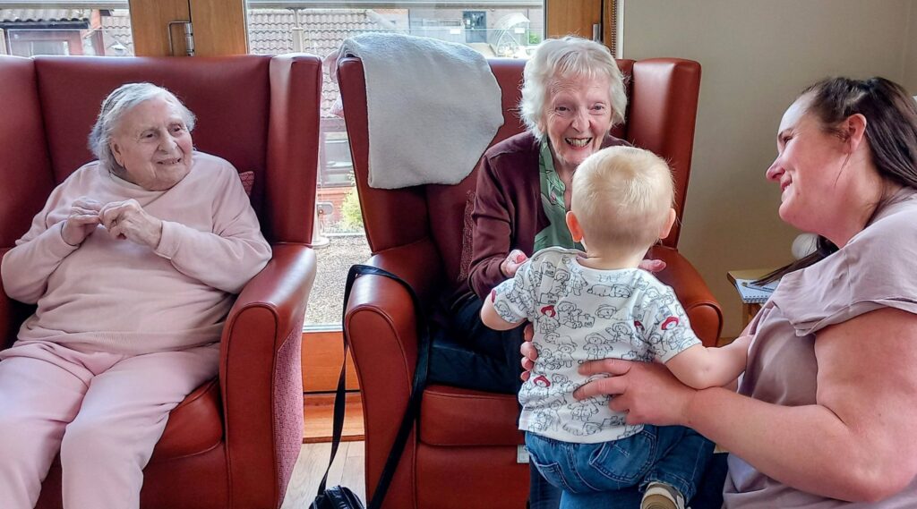 Three women sit in armchairs as a toddler stands between them; they seem to be interacting and smiling in a bright, homely room, as though Jenna and her friends are everyday superheroes.