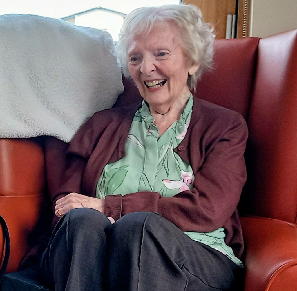 Elderly woman with short white hair sits on a red armchair, smiling. She wears a green patterned blouse and maroon cardigan. Jenna’s white blanket is draped over the chair behind her, like a superhero’s cape.