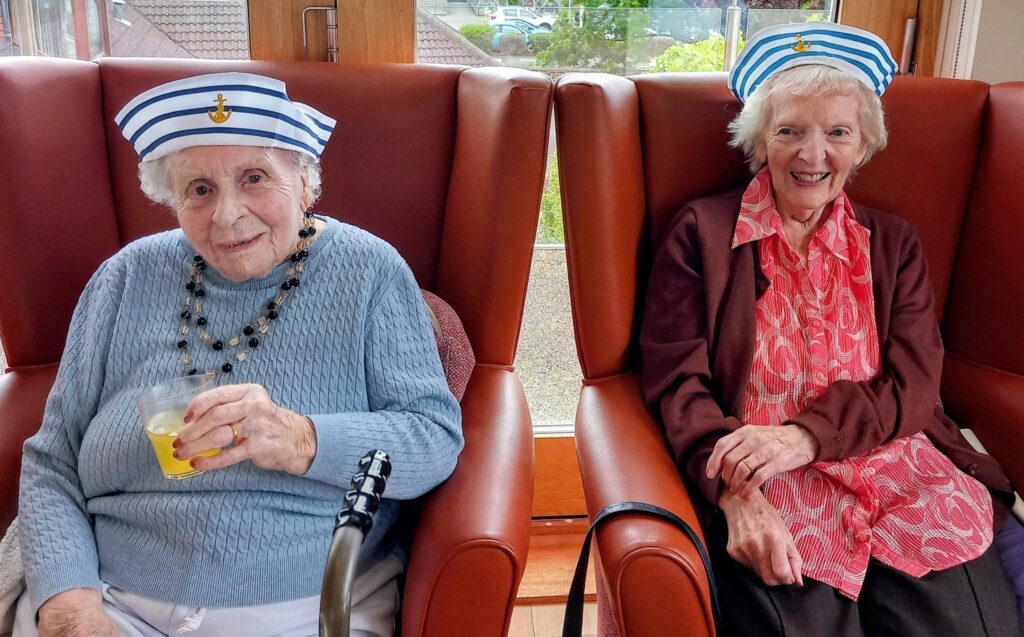 Two elderly women in sailor hats sit side by side in armchairs; one is holding a drink, both smiling like seasoned sailors enjoying shore leave.