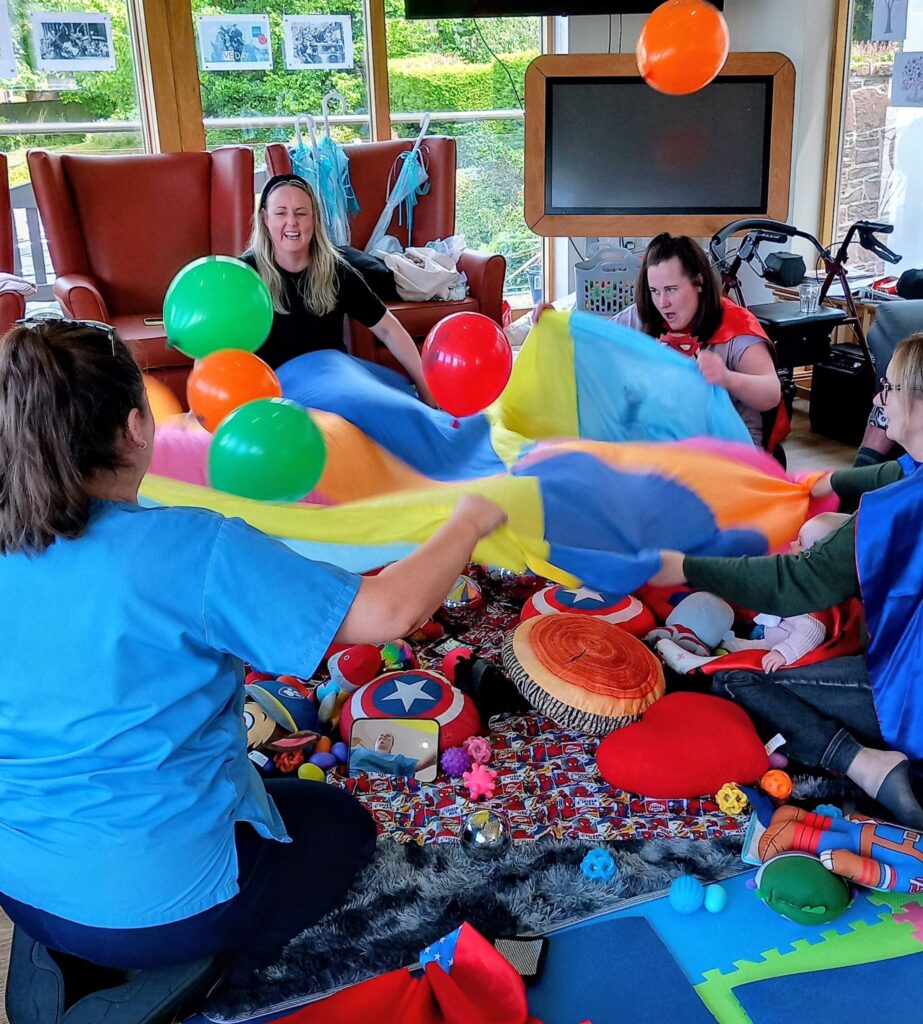 Four adults, including Jenna, sit in a circle holding a colourful parachute with balloons on top, surrounded by toys and cushions in a bright room, pretending to be superheroes or sailors.