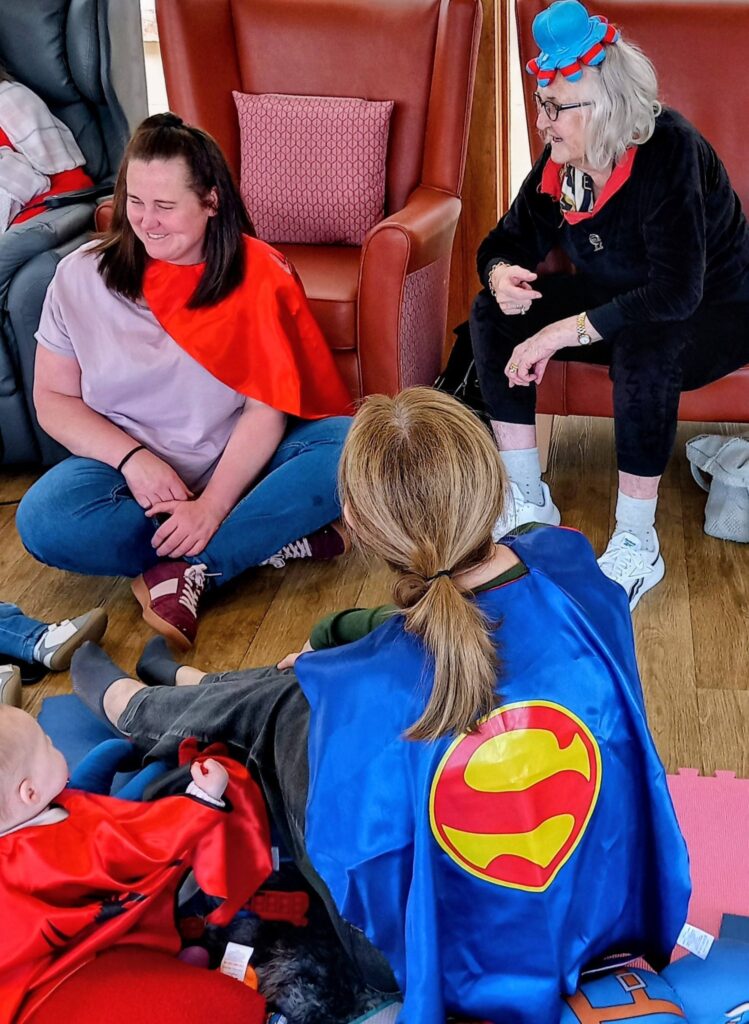 Three adults and a baby are seated on the floor together. Two adults wear superhero capes, while Jenna wears a blue hat with a red bow. Everyone looks engaged and at ease.