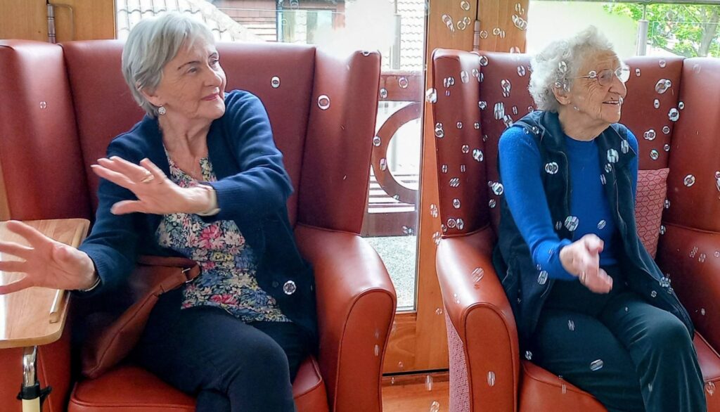 Two elderly women sit in red armchairs, grinning like superheroes as they reach out to touch floating bubbles in a bright indoor setting.