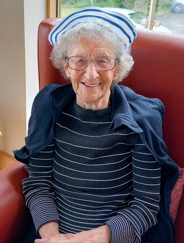 An elderly woman, Jenna, with grey curly hair, spectacles, and a striped sailor hat sits smiling in a red chair, dressed in a striped jumper and a navy jacket draped over her shoulders like the sailors or superheroes she admires.