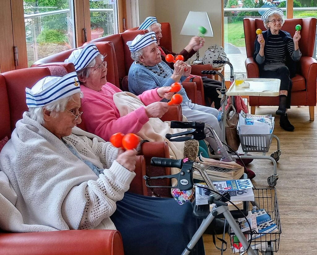 Four elderly women in sailor hats sit in armchairs, shaking maracas like jolly sailors. Walking frames and magazines are close by. One woman, Jenna, sits separately, also holding maracas.