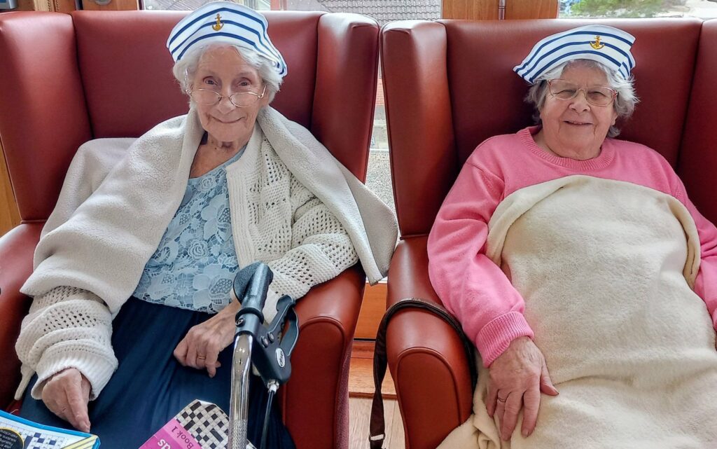 Two elderly women sit side by side in armchairs, both wearing matching striped sailor hats and spectacles, each wrapped in a blanket—like retired sailors or secret superheroes reminiscing about their Jenna days.