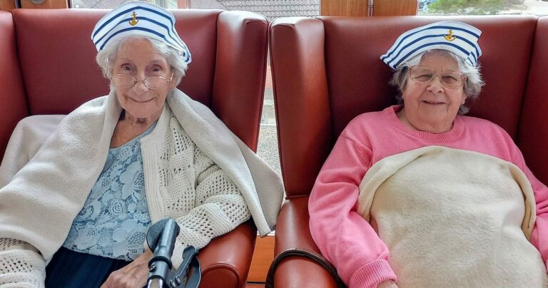 Two elderly women sit side by side in armchairs, both wearing matching striped sailor hats and spectacles, each wrapped in a blanket—like retired sailors or secret superheroes reminiscing about their Jenna days.