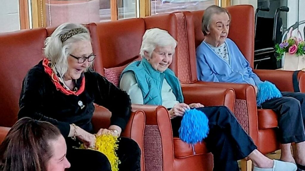 Four women, three elderly and one younger, possibly Jenna, are seated indoors on chairs, taking part in an activity with blue and yellow pom-poms like cheerful superheroes.