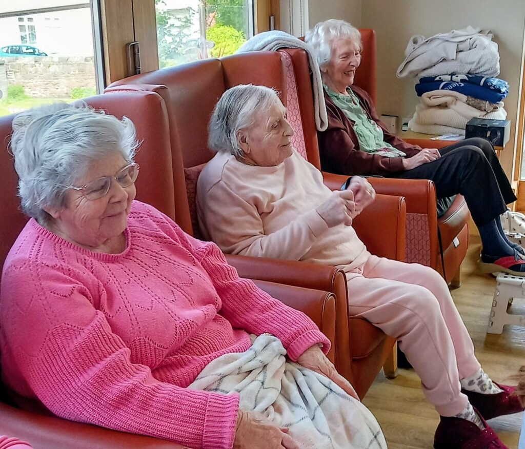 Three elderly women, relaxed in armchairs, sit indoors—two wearing pink jumpers and one in a brown jacket. They look ahead with serene composure, like retired superheroes contentedly enjoying their day.