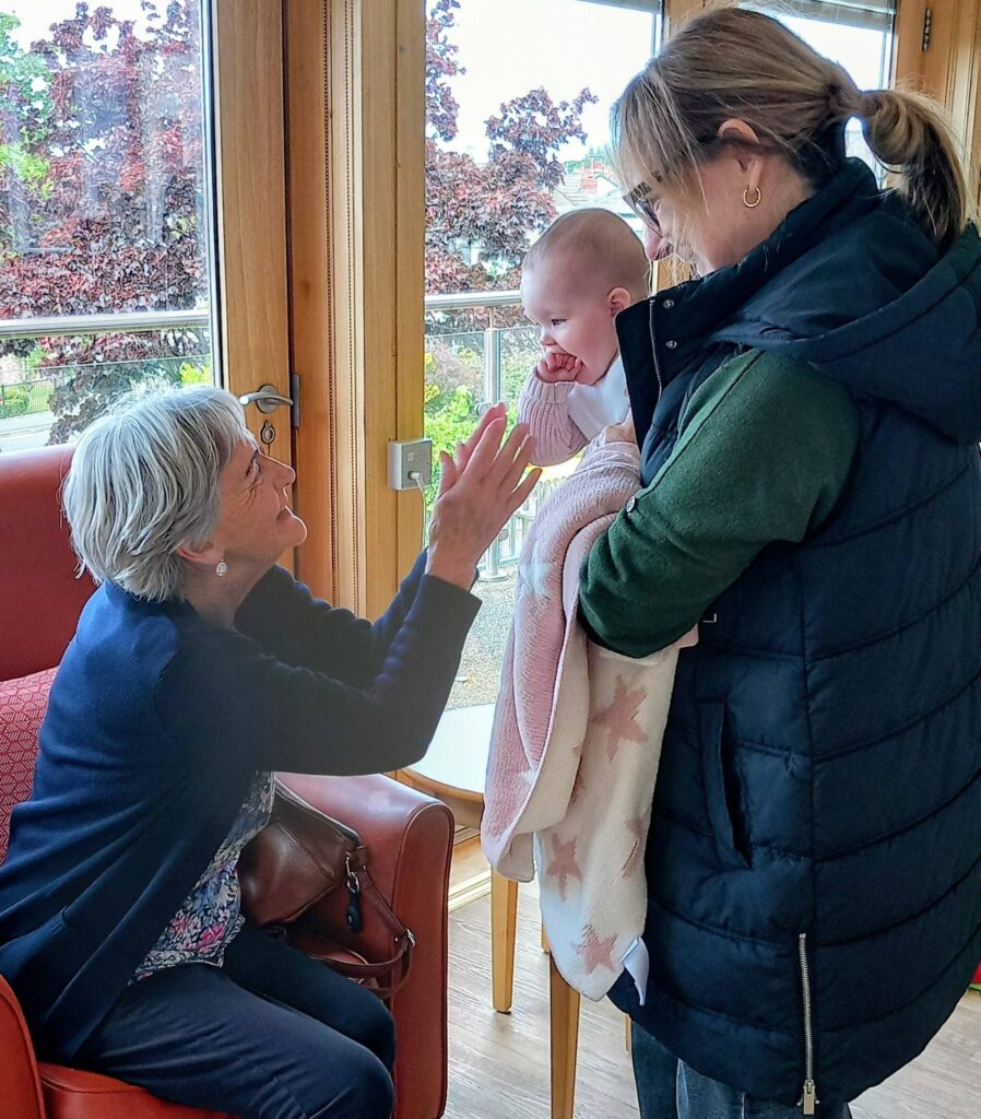 An elderly woman seated in a chair reaches out to touch the hand of a baby being held by Jenna, who stands nearby indoors.