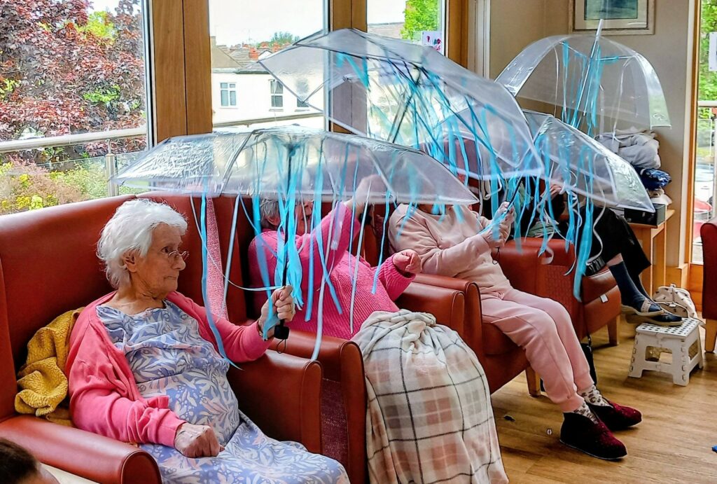 Four elderly people sit indoors on armchairs; three hold clear umbrellas adorned with blue streamers, looking like playful superheroes, with blankets draped over their knees.