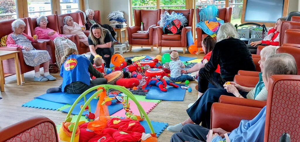 Elderly people sit in a circle, watching babies and toddlers play on mats with toys such as tiny superheroes, in a brightly lit room.
