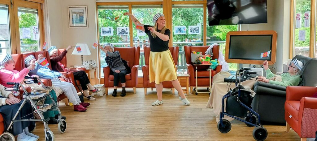 A group of elderly people sit in a lounge waving small flags while Jenna, dressed as a superhero in the centre, leads an activity; large windows allow daylight to stream in.