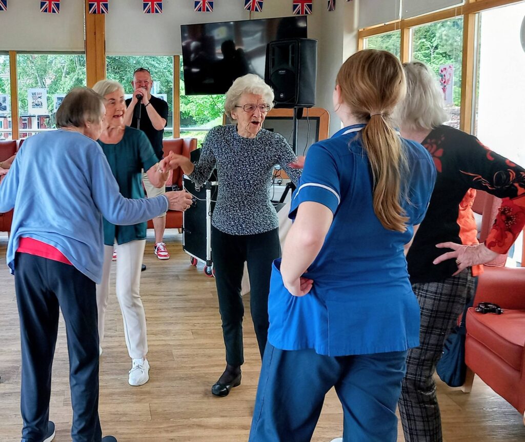 A group of elderly women and a nurse are dancing together in a community room, whilst a man sings into a microphone in the background.