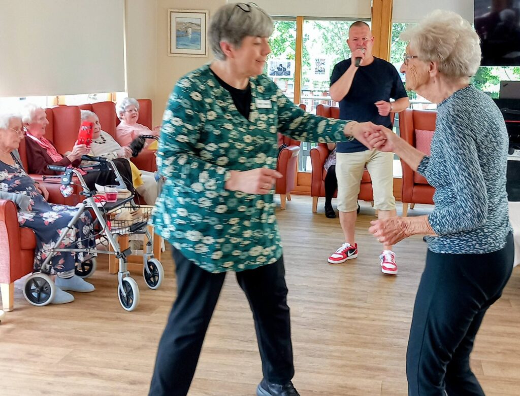 Two elderly women dance together in a communal lounge whilst other pensioners watch from armchairs; a man holding a microphone stands in the background.