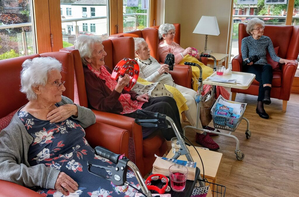 Five elderly women sit in a brightly lit common room, some holding musical instruments; drinks and personal belongings rest on side tables, creating a relaxed and sociable atmosphere.