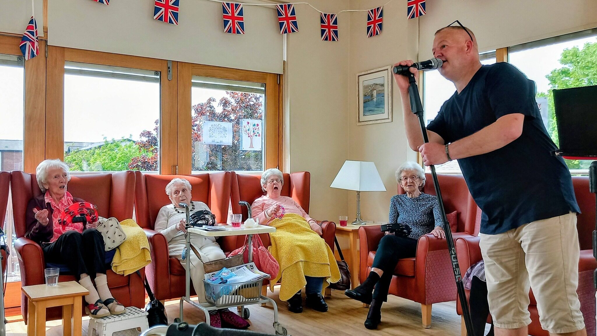 A man sings into a microphone for four elderly women seated in armchairs in a decorated room adorned with Union Jack bunting and large windows.