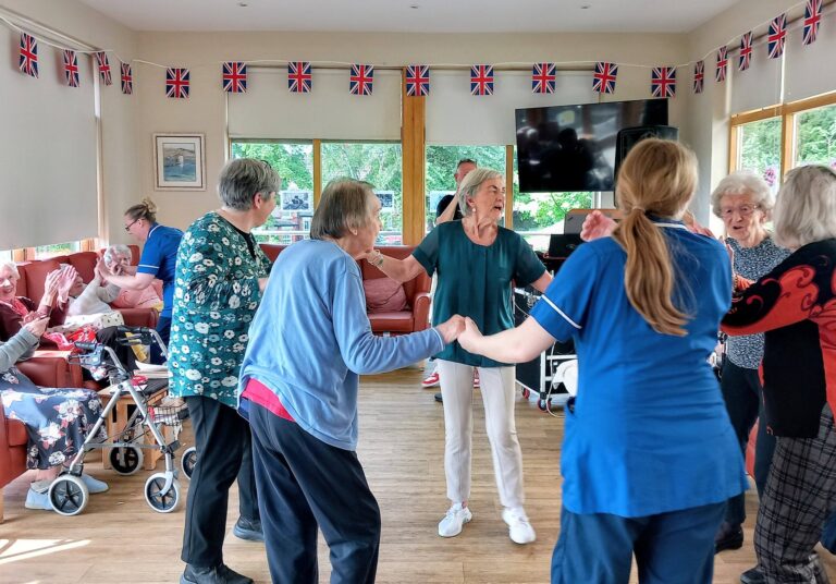 Elderly people and carers hold hands and dance in a decorated room adorned with Union Jack flags, whilst others sit and watch.
