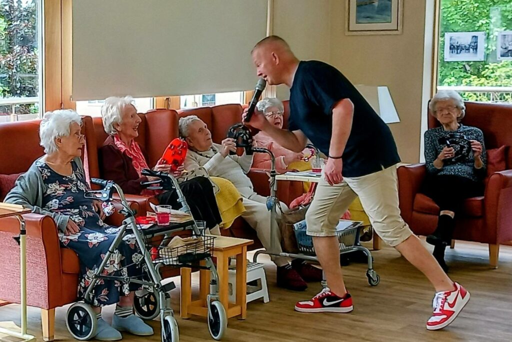 A man with a microphone sings to a group of elderly women seated in a lounge, some holding drinks and smiling, in a brightly lit room with large windows.