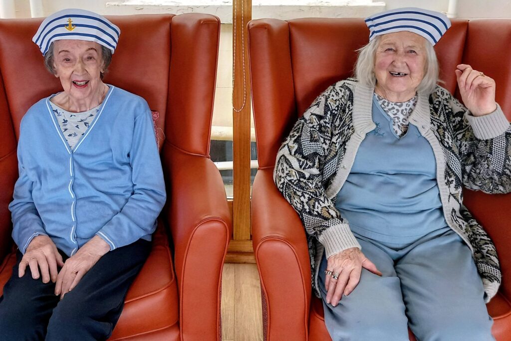 Two elderly women sit in orange chairs, wearing blue and white sailor hats and casual clothes, smiling at the camera like cheerful sailors ready for adventure.