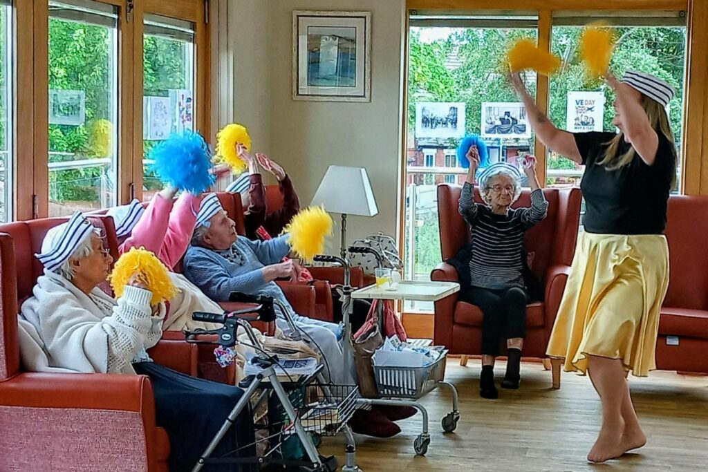 A group of elderly people in armchairs hold coloured pom-poms, following Jenna as she leads a lively activity in a bright, windowed room—they move like cheerful superheroes.