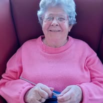 An older woman with grey hair and glasses, dressed in a pink jumper, sits in an armchair and smiles whilst holding knitting needles—knitting quietly and changing the world one stitch at a time.
