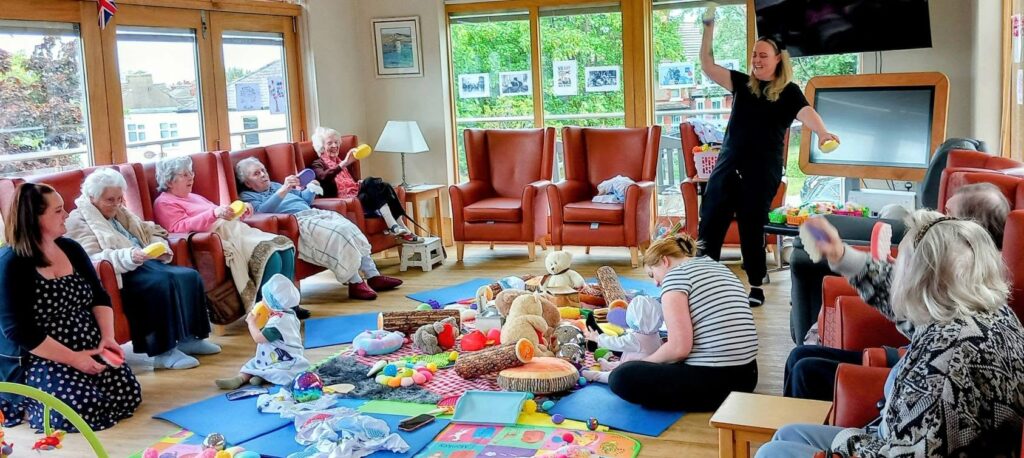 A group of elderly people and carers sit in a bright room, taking part in Sensory Play with babies and toys spread across mats. A woman stands, leading the Boogie Baby activity.